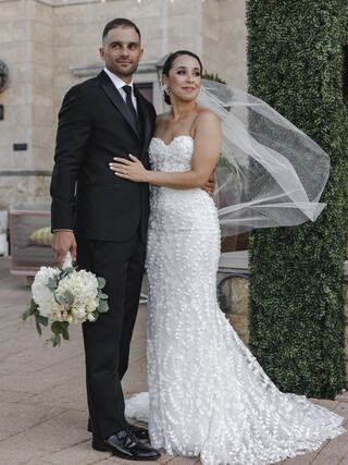 A couple stands together, dressed elegantly for a wedding, with the bride in a stunning white gown and the groom in a sharp black suit.