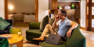 A man sits comfortably on a green couch, talking on the phone with a laptop beside him, in a modern, well-lit lounge area.