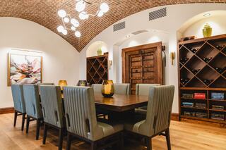 A stylish dining area with a large wooden table, plush chairs, a decorative light fixture, and wine storage against the walls.