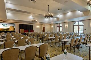 A spacious conference room with rows of tables and chairs, featuring large windows, chandeliers, and a backdrop displaying "Hotel Viala."
