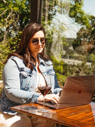 A woman in sunglasses works on a laptop at a table outdoors, with a drink beside her, surrounded by greenery and sunlight.