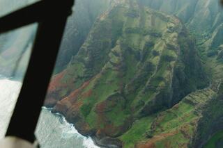 Lush, green cliffs rise dramatically above a coastal shoreline, with waves crashing against rocks beneath a misty, serene sky.