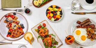 A vibrant brunch spread featuring waffles with berries, avocado toast, fried eggs, sausages, fresh fruit, and refreshing beverages.