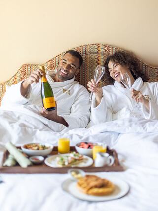 A joyful couple in bathrobes enjoy champagne and breakfast in bed, smiling and celebrating together. Cozy and intimate morning vibes.
