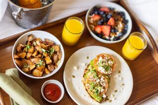 A tray presents a delightful breakfast: avocado toast, roasted potatoes, a mixed fruit bowl, and refreshing orange juice.