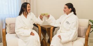Two women in white bathrobes happily toast with glasses of green drinks while sitting in cozy chairs in a bright room.