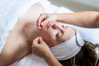 A person receives a facial massage, eyes closed, relaxed on a treatment table, wearing a headband and surrounded by soft linens.
