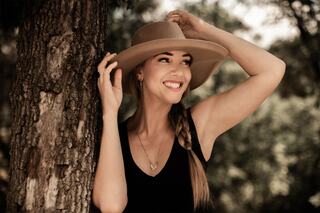 A smiling woman with a stylish hat playfully poses beside a tree, exuding charm and confidence in a soft, natural setting.