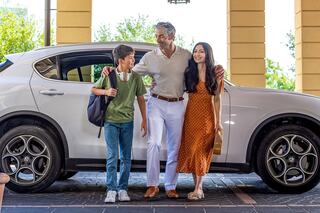 A man, woman, and boy share a joyful moment outside a white car, with trees in the background, embodying a sense of family and togetherness.
