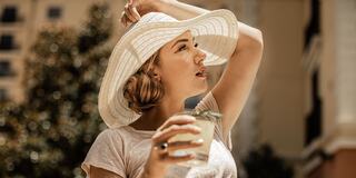 A woman in a wide-brimmed hat enjoys a drink, looking thoughtfully into the distance against a sunny backdrop.
