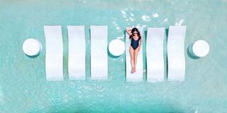 A woman relaxes on a white lounge in a turquoise pool, surrounded by modern, minimalist curves and circular tables. Sunlight glistens on the water.