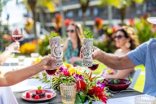 Two people toast with tiki mugs at a vibrant outdoor gathering, surrounded by tropical flowers and cheerful friends enjoying drinks.