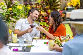 A couple toasts joyfully at a festive outdoor meal, surrounded by vibrant plants and floral decorations.
