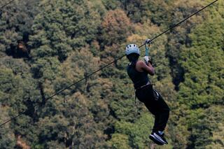A person descends a zip line, wearing a helmet and sporty attire, with a backdrop of lush green trees and hills. Excitement in the air!