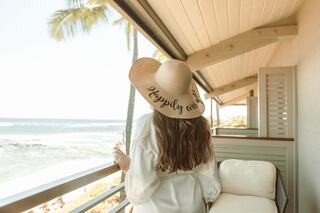 A woman in a large sun hat enjoys a scenic ocean view from a balcony, radiating a relaxed, carefree vibe.