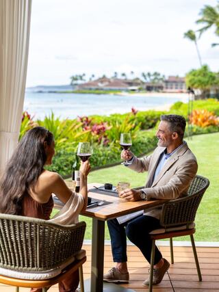 A couple toasts with drinks at a scenic table overlooking lush greenery and the ocean, enjoying a relaxed moment together.