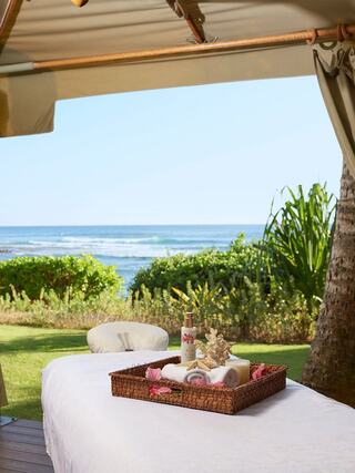 A serene outdoor spa setup overlooks the ocean, featuring a massage table, a woven basket with towels, and tropical greenery.