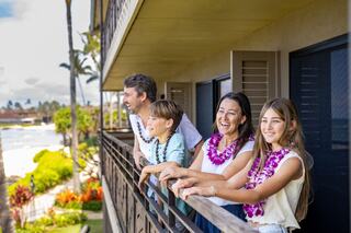 A cheerful family stands on a balcony, wearing leis and enjoying the scenic view of the ocean and vibrant greenery.