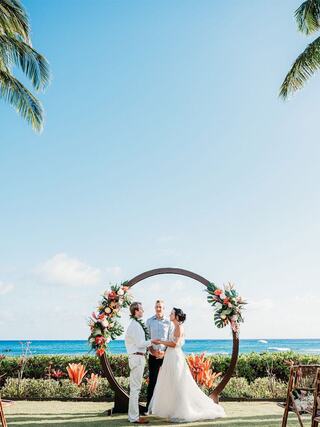 A couple exchanges vows under a floral arch by the ocean, surrounded by tropical plants and bright blue skies. A joyful moment unfolds.