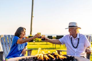 Two people toast with drinks while seated by a fire, enjoying a sunny outdoor setting with a serene backdrop.