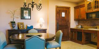 A cozy dining area features a round table with blue chairs, a wooden cabinet, and a well-equipped kitchen, complemented by soft lighting.