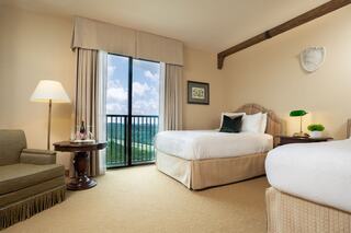 A cozy hotel room features two beds, a seating area, a large window with a view, and warm decor, creating an inviting atmosphere.