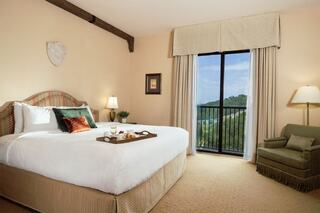 A cozy hotel room featuring a large bed, a small table with breakfast, a comfy chair, and a view through glass doors.