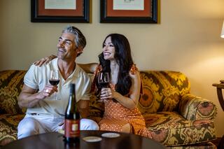 A couple enjoys a cozy moment on a floral-patterned couch, laughing and sipping red wine together, radiating joy and connection.