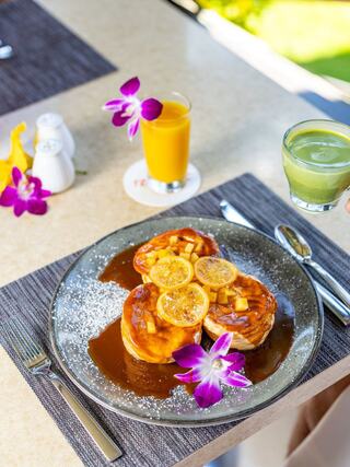 Fluffy pancakes topped with caramelized citrus and edible flowers, accompanied by a vibrant juice and green drink on a stylish table setting.