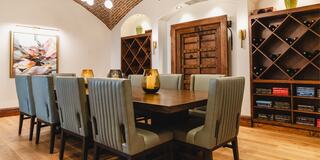 A cozy dining area features a wooden table, elegant chairs, an art piece, and wine storage, accentuated by warm lighting and rustic architecture.