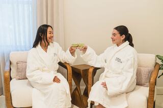 Two women in matching white robes toast with drinks, enjoying a relaxed moment together in a cozy setting.