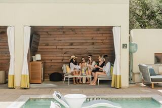A group of five friends relaxes by a pool, enjoying drinks and conversation in a stylish cabana with a cozy atmosphere.