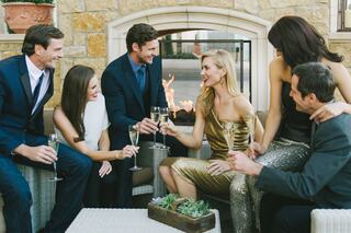 A stylish group of well-dressed friends celebrates together, toasting with champagne in a cozy setting by a fire. Laughter and joy abound.