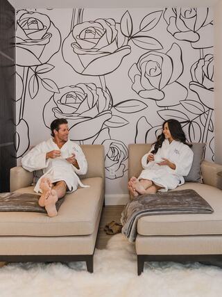 A couple in white robes relaxes on cozy lounges, enjoying snacks, with a floral mural as a backdrop, exuding a soothing ambiance.