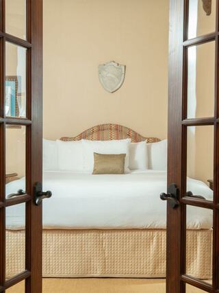 A cozy bedroom features a large bed with decorative pillows, framed by elegant wooden doors. Earthy tones create a warm, inviting atmosphere.