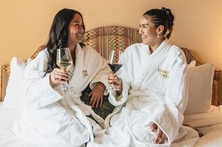 Two women in plush white robes enjoy drinks while sitting together on a bed, sharing smiles and laughter.