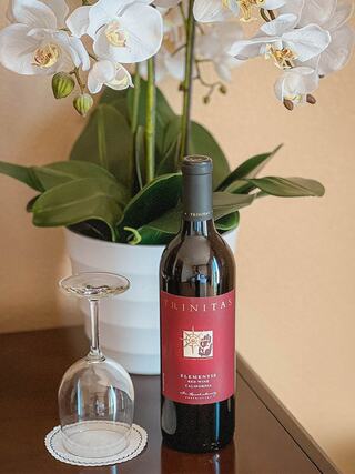 A bottle of Trinitas wine sits beside a clear wine glass on a polished table, accompanied by elegant white orchids in a pot.