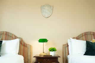 A cozy, well-decorated bedroom features two beds with patterned headboards, a green lamp on a nightstand, and a decorative wall shield.