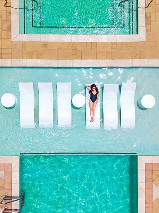 A woman relaxes on a sun lounger in a bright pool, surrounded by inviting turquoise water and sleek white lounge chairs.