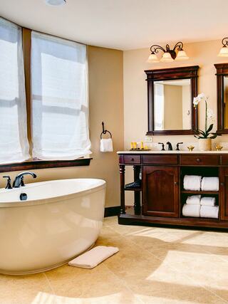 A bright, elegant bathroom features a freestanding tub, wooden vanity with towels, and natural light from large windows.