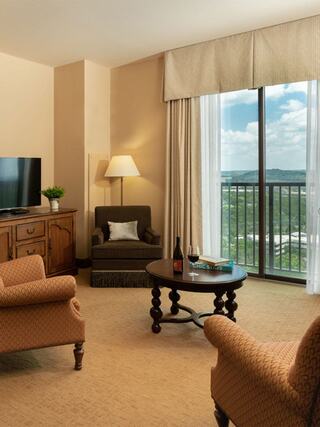 A cozy hotel room features two armchairs, a wooden table with wine, a TV, and large windows revealing a scenic view and soft natural light.