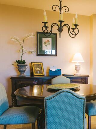 A cozy dining area featuring a round table, upholstered chairs, elegant chandelier, and decor with a plant and framed artwork.