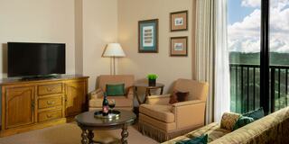 A cozy living area features a TV, comfortable armchairs, and a small table with drinks, illuminated by natural light from large windows.