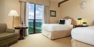 A cozy hotel room with two beds, a chair, a lamp, and a large window showcasing a scenic view of green hills and a blue sky.