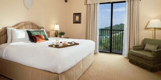 A cozy hotel room features a king-sized bed, a small dining tray, and a balcony with scenic views, complemented by soft lighting and elegant decor.