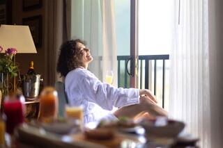 A woman in a robe relaxes by a light-filled window, enjoying a drink, surrounded by a spread of breakfast items and flowers.