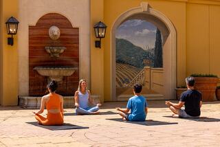 A serene outdoor yoga session with four participants in meditation, set against a beautiful mural and fountain backdrop.
