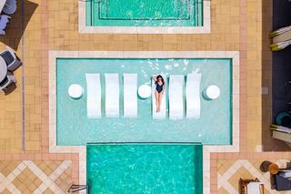 A person relaxes on pool loungers in a vibrant, turquoise pool area surrounded by sunbathers and stylish furniture.