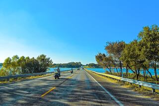 A scenic road stretches ahead, flanked by lush trees and a shimmering body of water, inviting a leisurely ride under a clear blue sky.