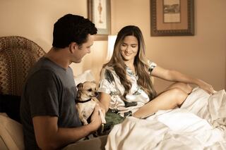 A couple sits on a bed, smiling and enjoying each other's company, with a small dog in their lap and soft bedding around them.